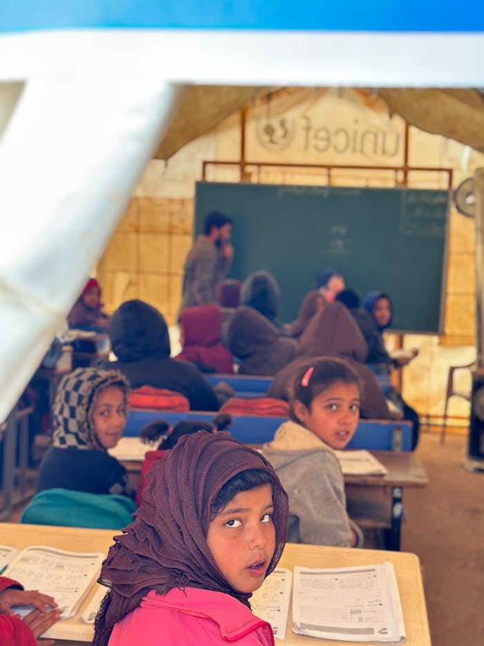 Children engaging in education inside a temporary classroom with a teacher and blackboard.