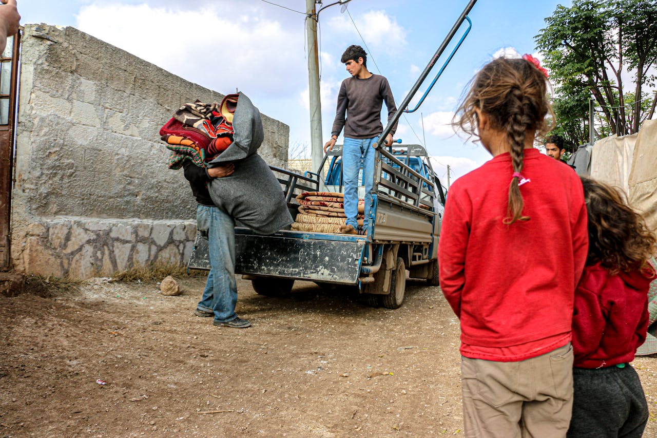 Anonymous man carrying bag of rugs between ethnic friends and children on dry roadway with truck under cloudy blue sky